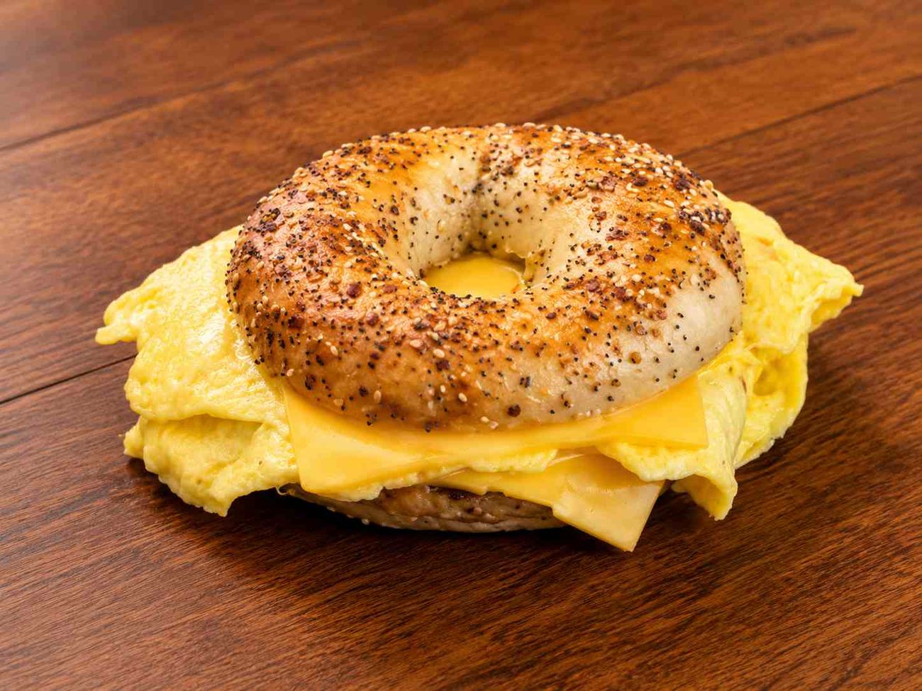 An everything bagel holding scrambled egg, melted cheese and a sausage patty, on a dark wood table.