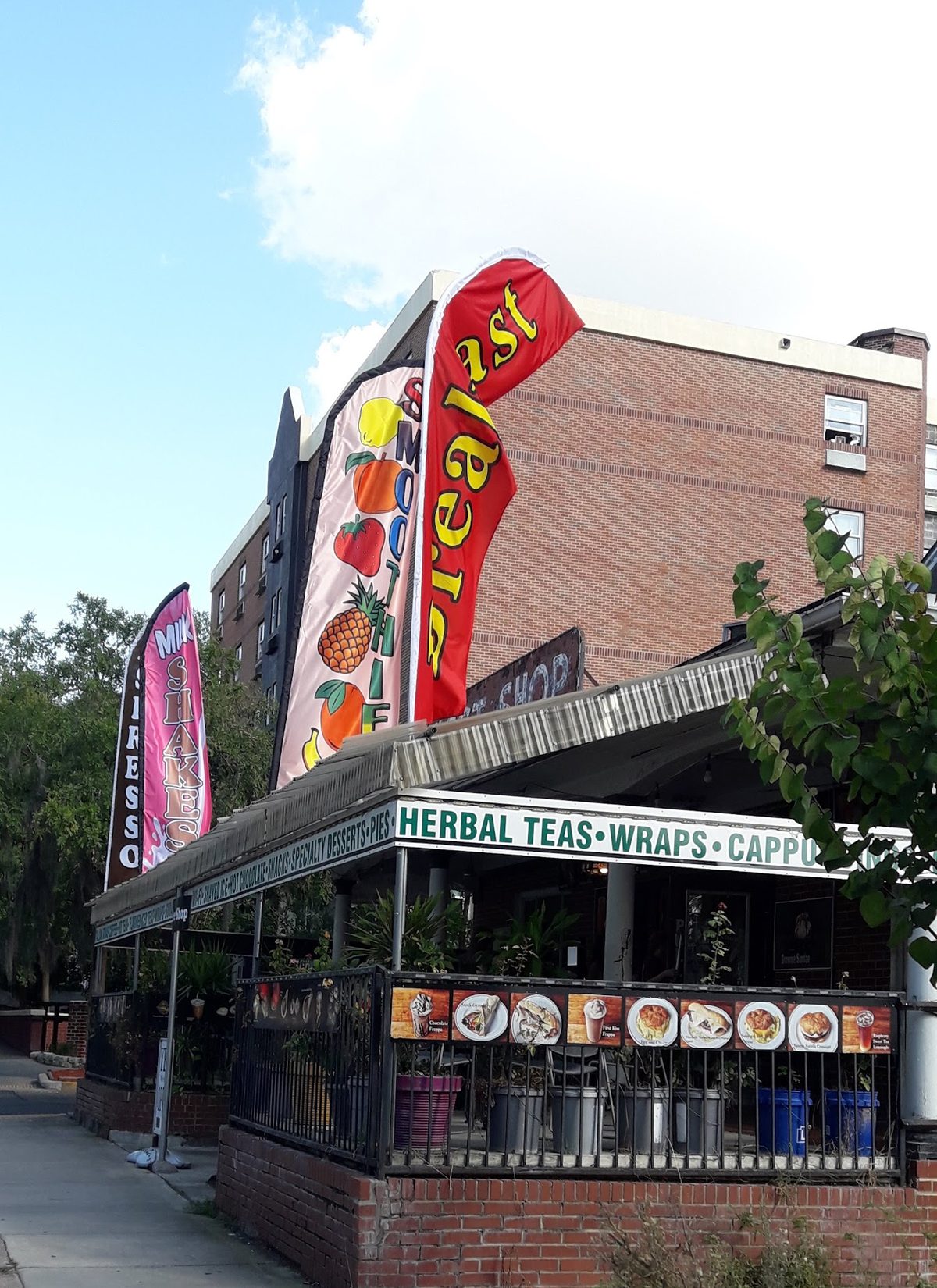 The corner shop under a metal awning, with a green and white sign board reading Herbal Teas, Wraps, Cappuccino and four tall flags on the sidewalk.